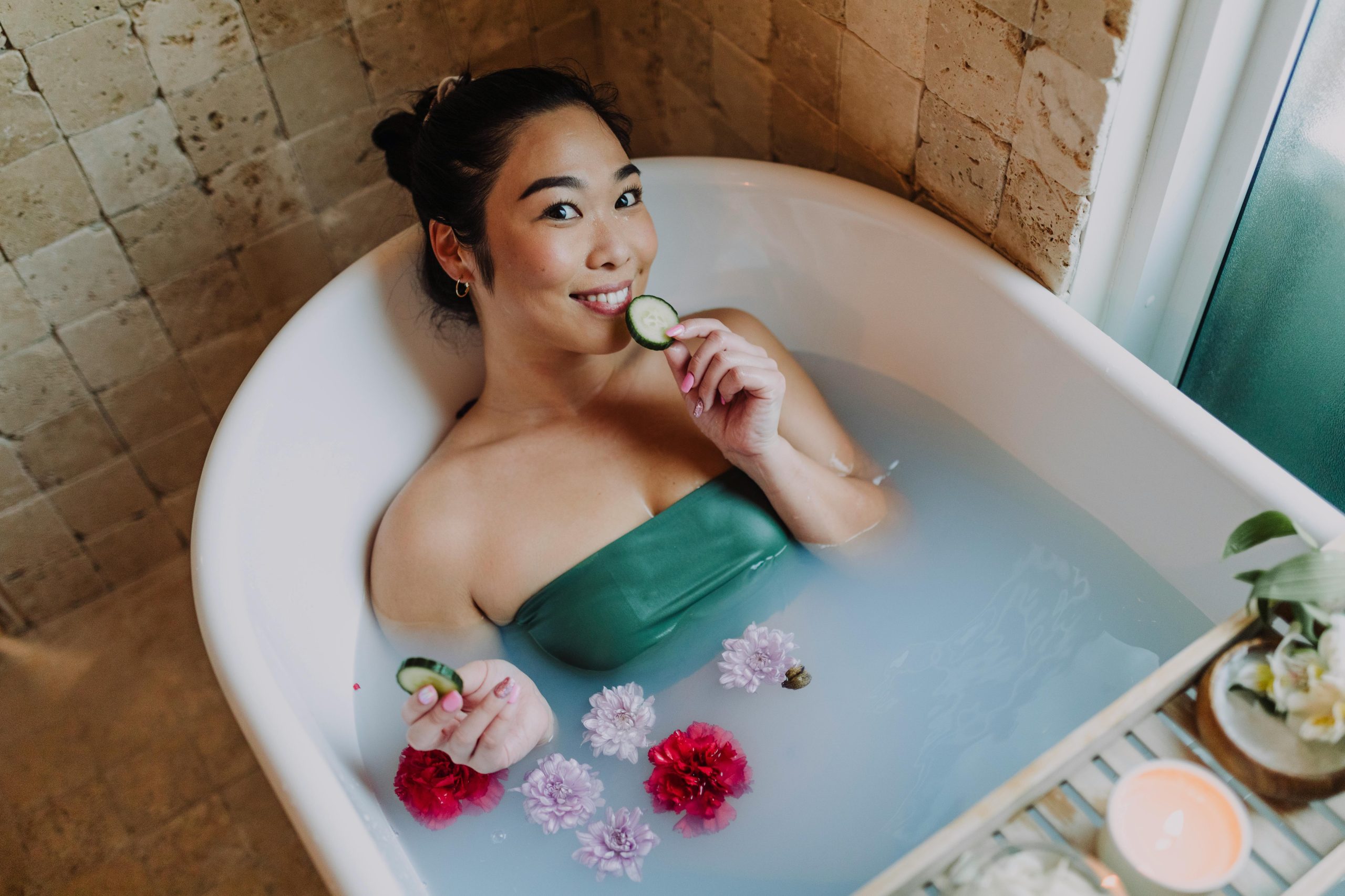 Asian woman enjoying a relaxing spa day at home in a flower-filled bath with cucumber slices.
