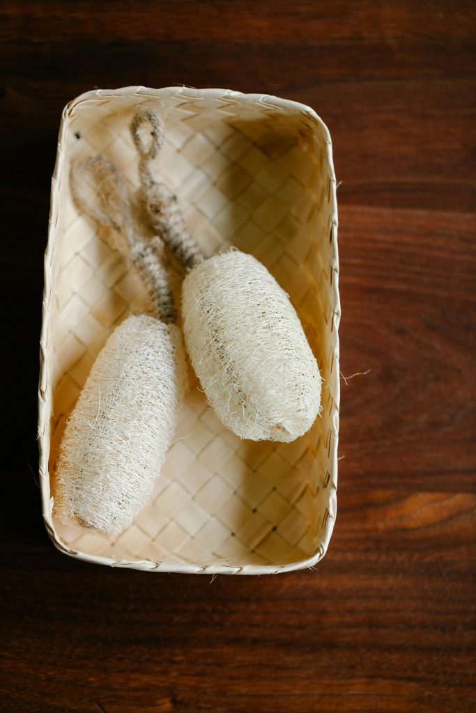 Two natural loofah sponges in a woven basket on a wooden surface.