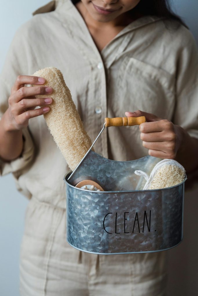 A woman holding a metal bucket filled with natural loofah and personal care items, emphasizing cleanliness.